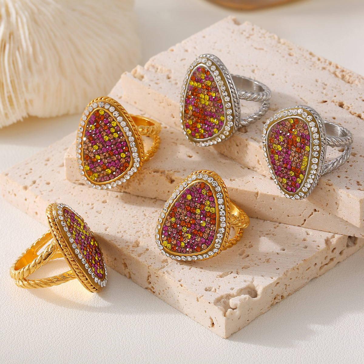 Gold-tone and steel-tone teardrop rings with multicolor stone faces arranged on pale stone display blocks.