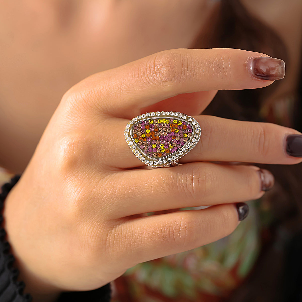 Close-up of a gold-tone teardrop cocktail ring with colorful stones worn on a hand with dark burgundy nails.