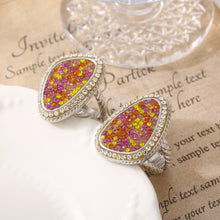 Two steel-tone teardrop cocktail rings with colorful stone clusters displayed on a white surface over a printed page.