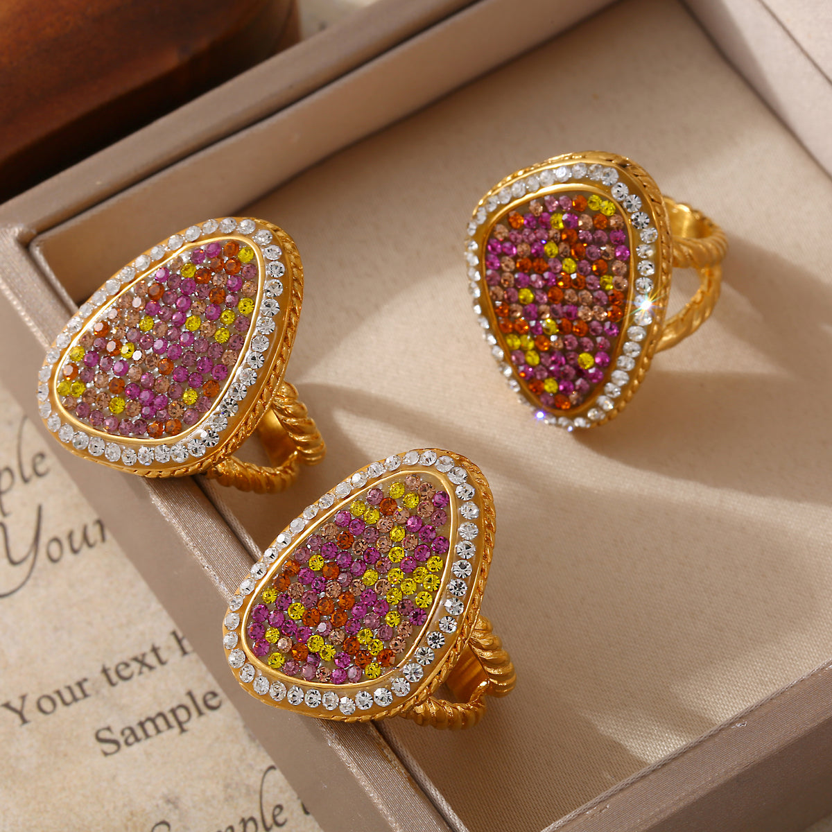 Three gold-tone teardrop statement rings with pink and yellow stone clusters arranged inside a beige jewelry tray.