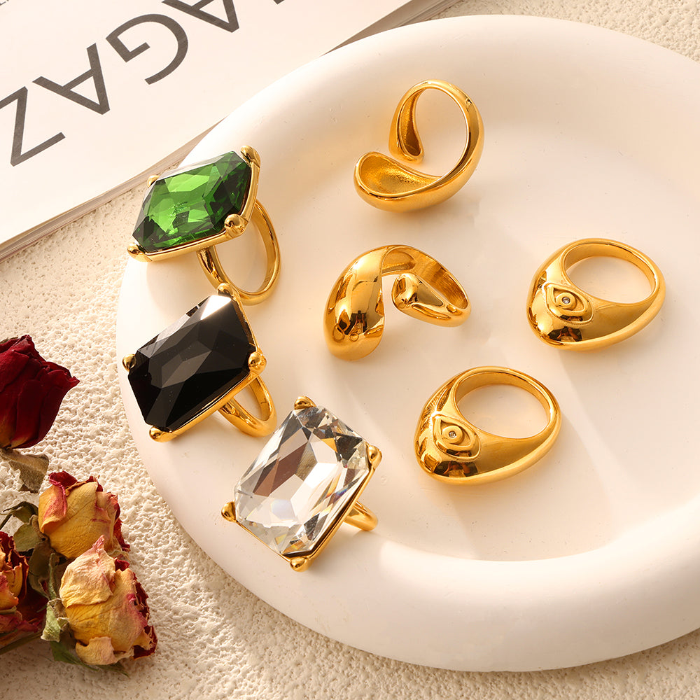 Gold-tone sculptural rings and clear, black and green rectangular stone rings arranged on a white tray