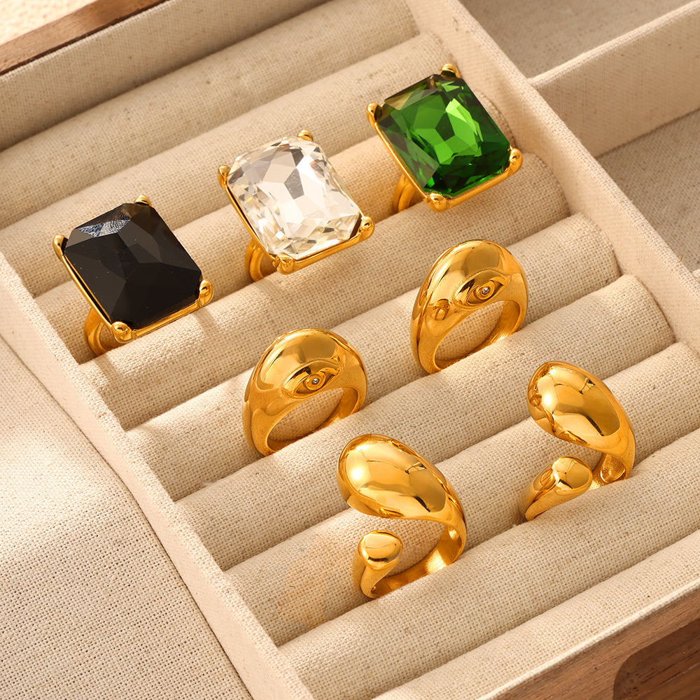 Clear, black and green rectangular stone rings displayed with gold-tone sculptural rings in a jewelry box