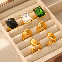 Clear, black and green rectangular stone rings displayed with gold-tone sculptural rings in a jewelry box