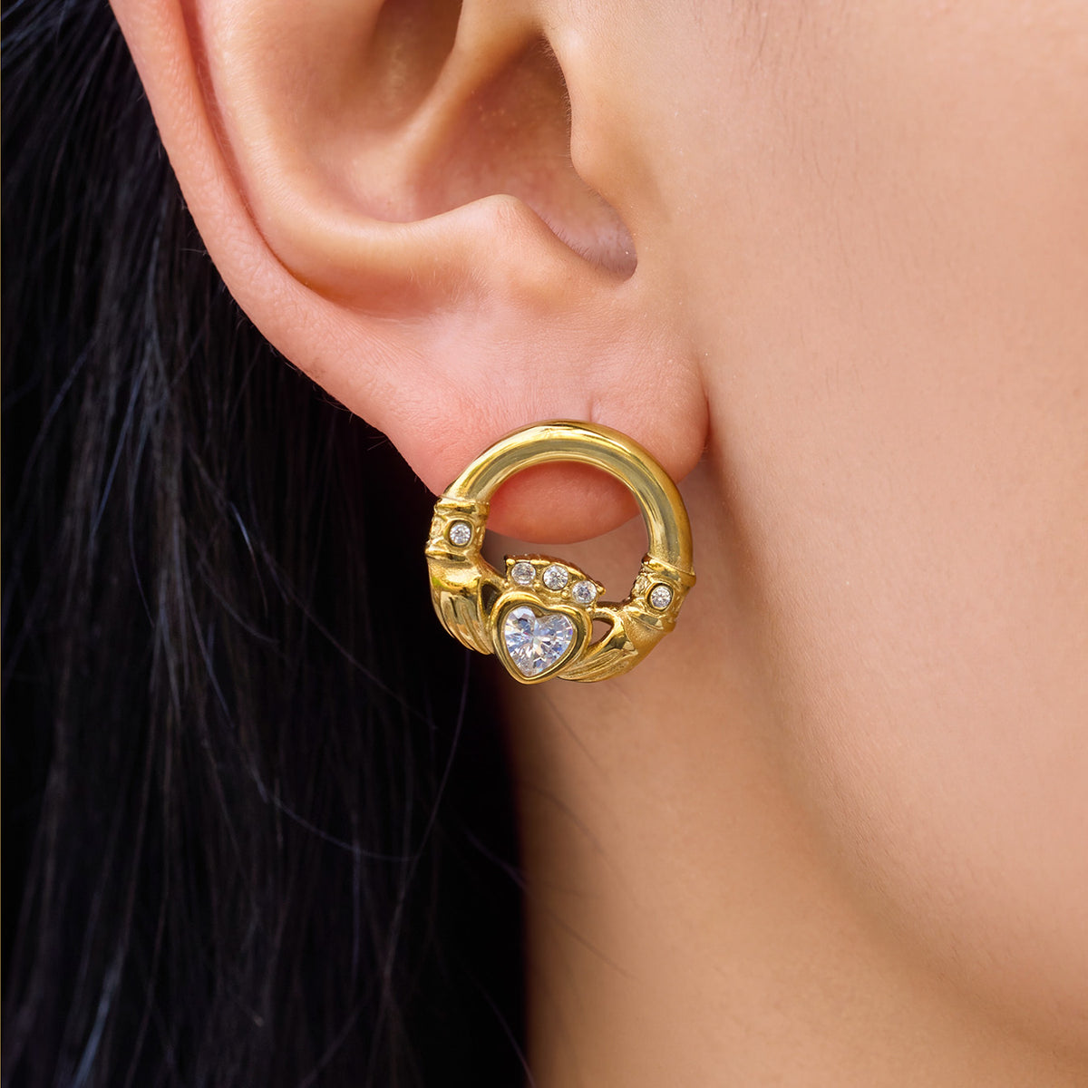 Close-up of a gold Claddagh-inspired heart earring on a model's ear, showing the hands and crowned heart motif