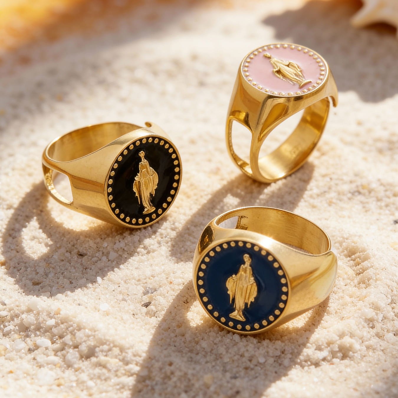 Color enamel Virgin Mary signet rings in gold stainless steel, black blue and pink styles displayed on sand