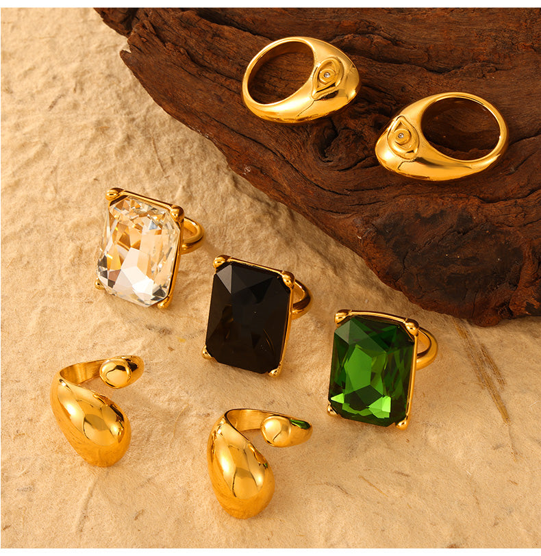 Clear, black and green rectangular stone rings with gold-tone sculptural rings on a textured background
