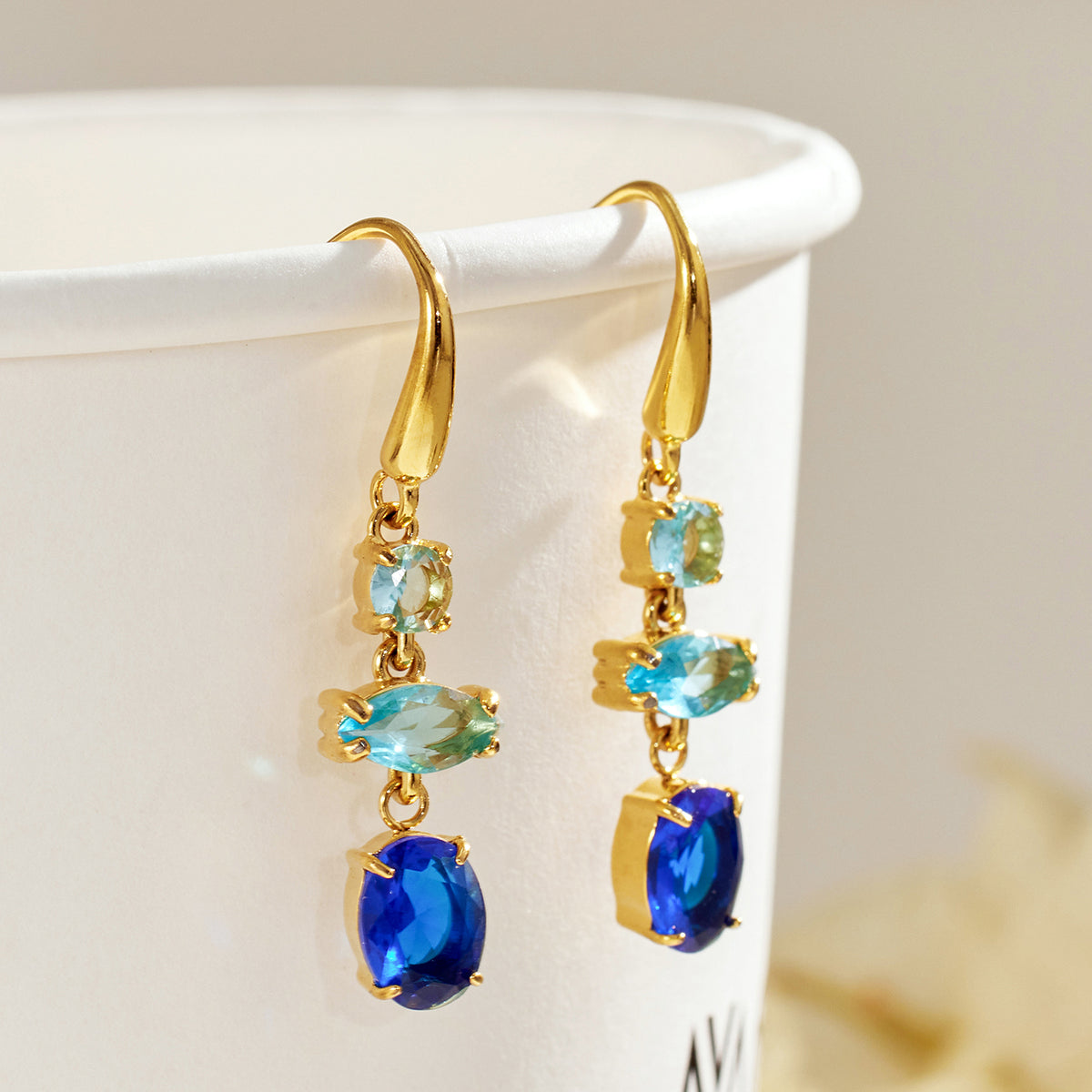 Pair of gold fishhook drop earrings with light blue marquise-shaped accents and deep blue oval stones hanging from a white display