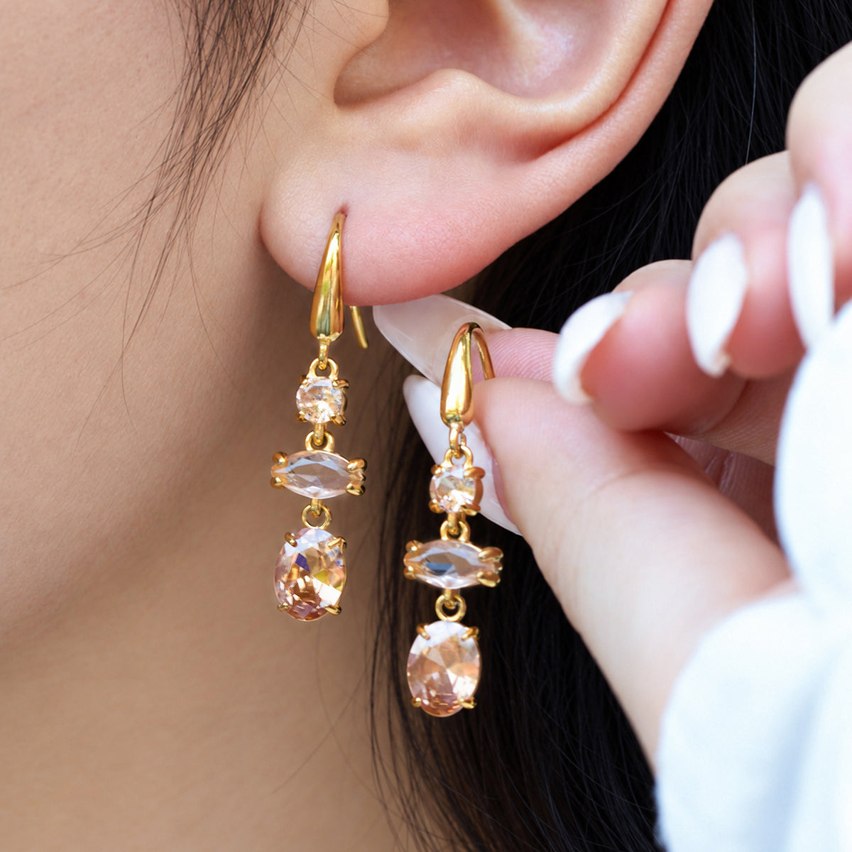 Model wearing champagne-colored three-stone drop earrings with gold fishhook wires while holding a matching earring beside her ear
