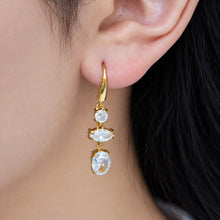 Model wearing a gold-tone three-stone drop earring with sparkling clear stones and a delicate oval pendant