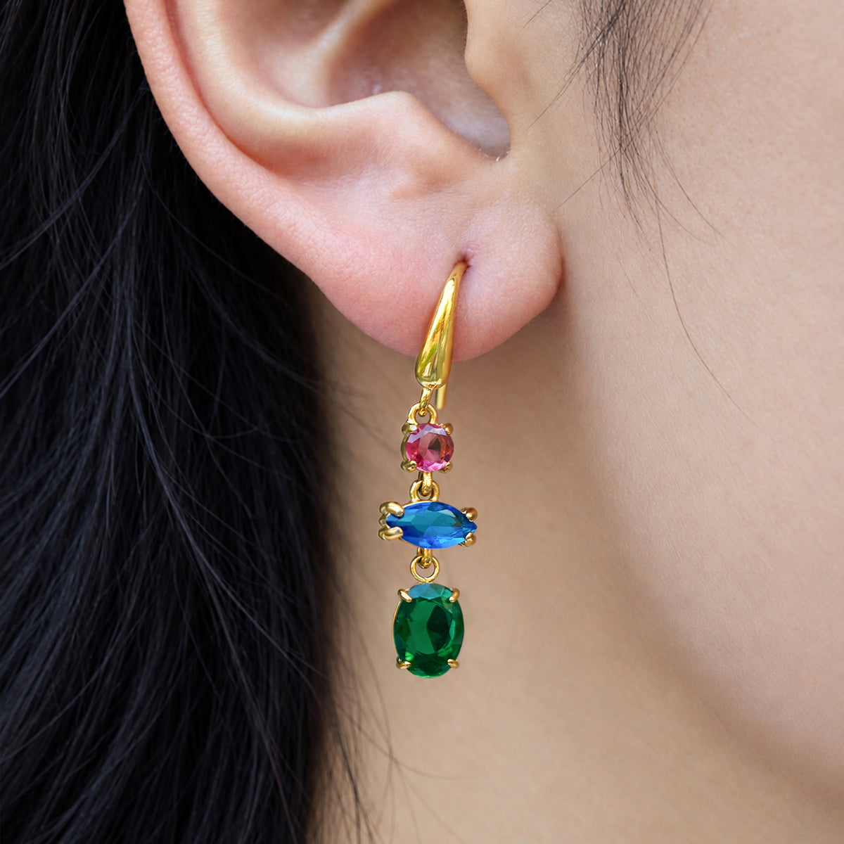 Close-up of a gold fishhook earring with pink, blue and green faceted stones hanging vertically from a model's earlobe