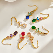 Flat lay of gold-tone stainless steel drop earrings with blue, red, green, purple, champagne and clear stones arranged on a light surface