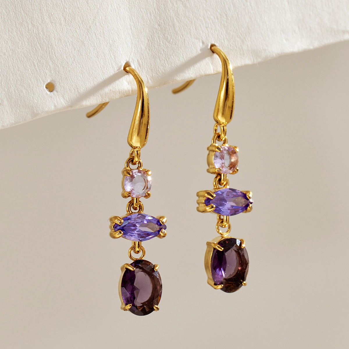Pair of gold-tone fishhook earrings with lavender and deep purple stones displayed against a neutral textured background