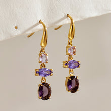 Pair of gold-tone fishhook earrings with lavender and deep purple stones displayed against a neutral textured background
