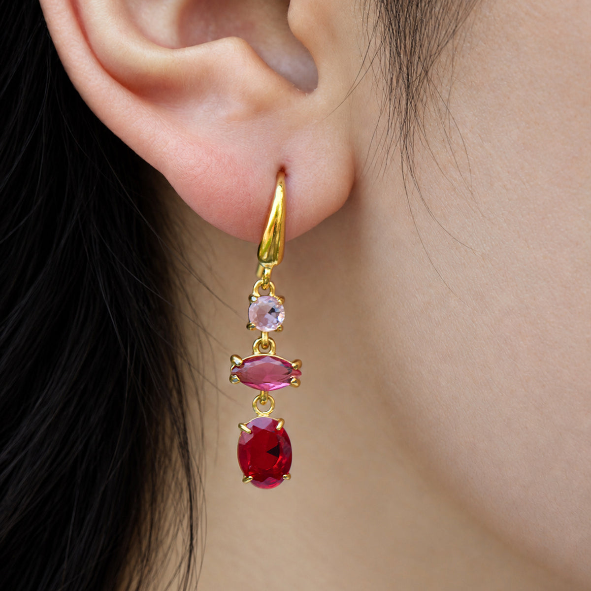 Gold-tone dangle earring with pink and red faceted stones worn on a model's ear, featuring a curved fishhook wire