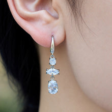 Model wearing a silver-tone stainless steel drop earring featuring three clear stones and an oval dangling accent