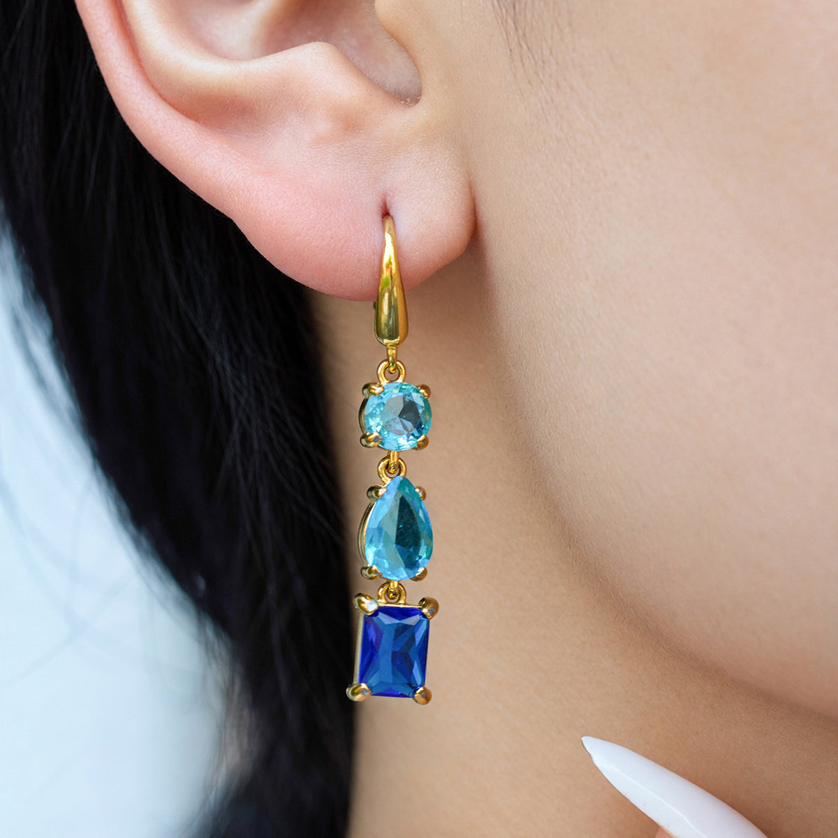 Model wearing a gold-tone geometric drop earring with aqua-blue upper stones and a deep blue rectangular bottom stone