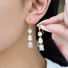 Model wearing clear three-stone gold drop earrings while holding a matching earring beside her ear to show the geometric design