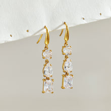Pair of gold fishhook earrings with clear round, pear-shaped and rectangular stones hanging against a light background