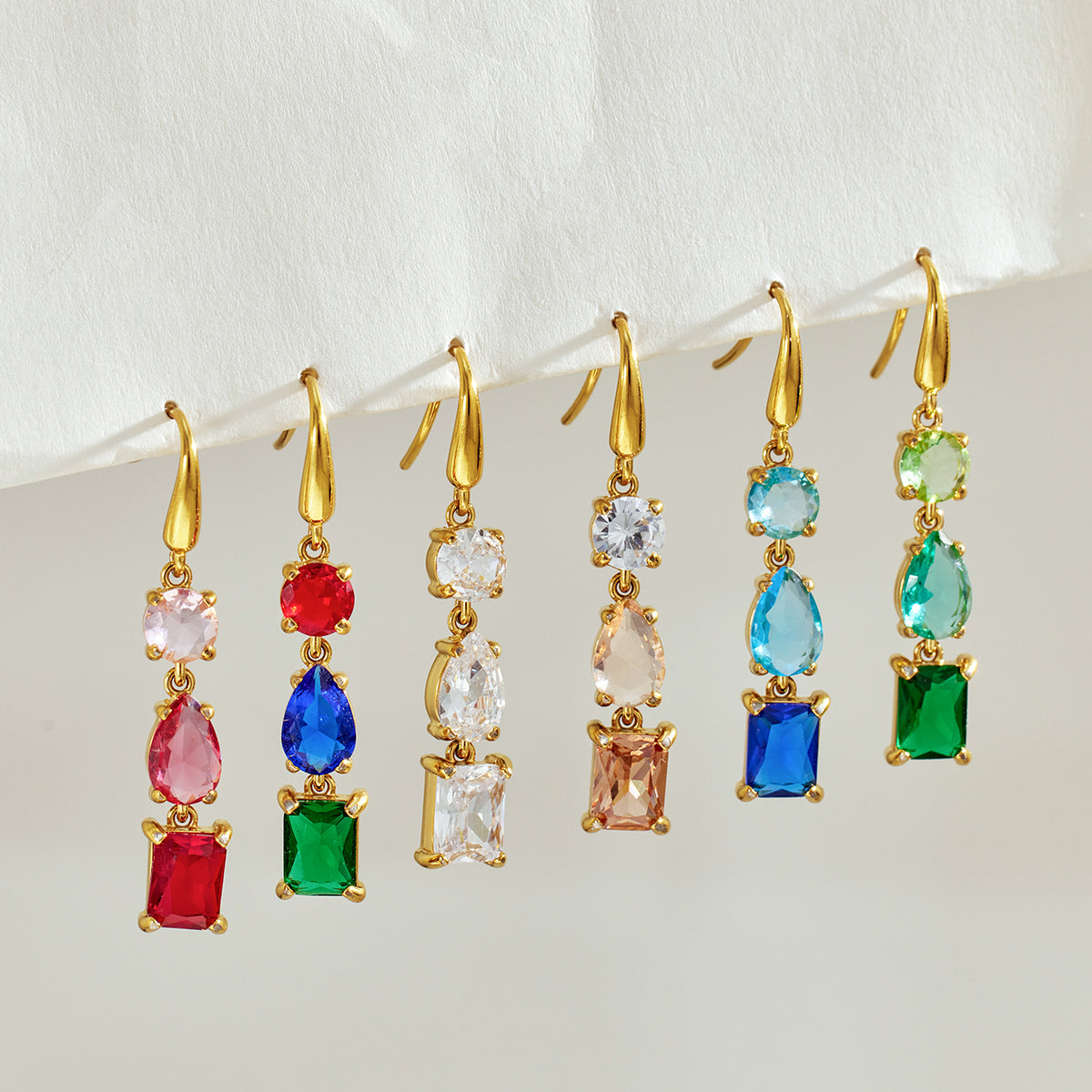Multiple gold-tone three-stone drop earrings hanging in a row, showing different faceted stone colors and geometric shapes