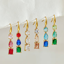 Multiple gold-tone three-stone drop earrings hanging in a row, showing different faceted stone colors and geometric shapes