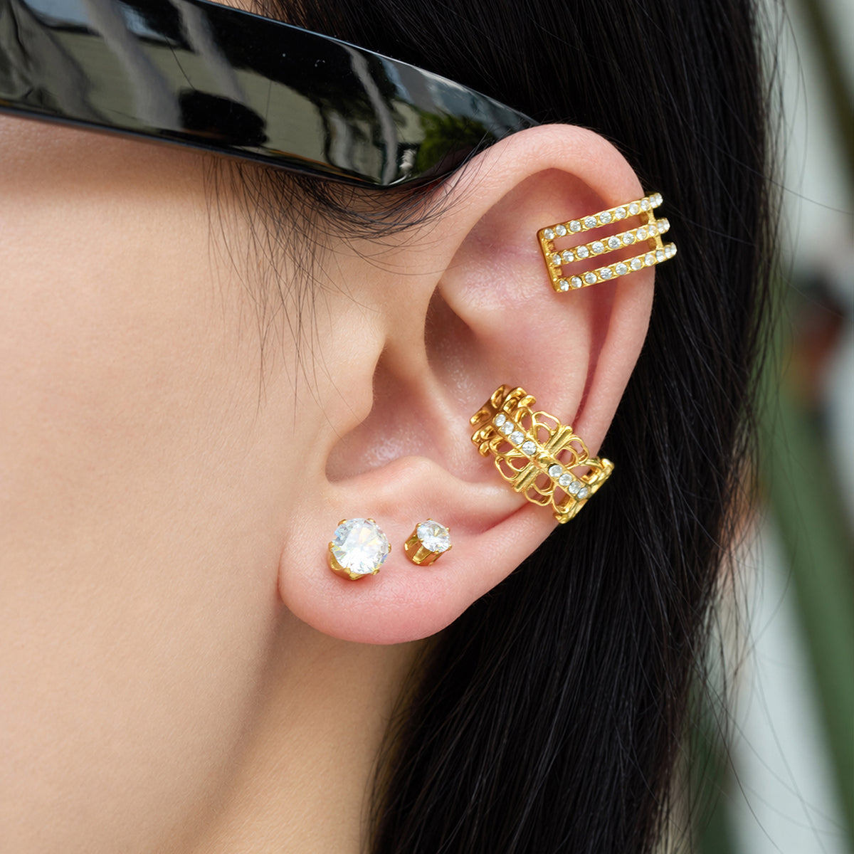Model wearing a small round clear stone stud earring styled with multiple gold ear accessories