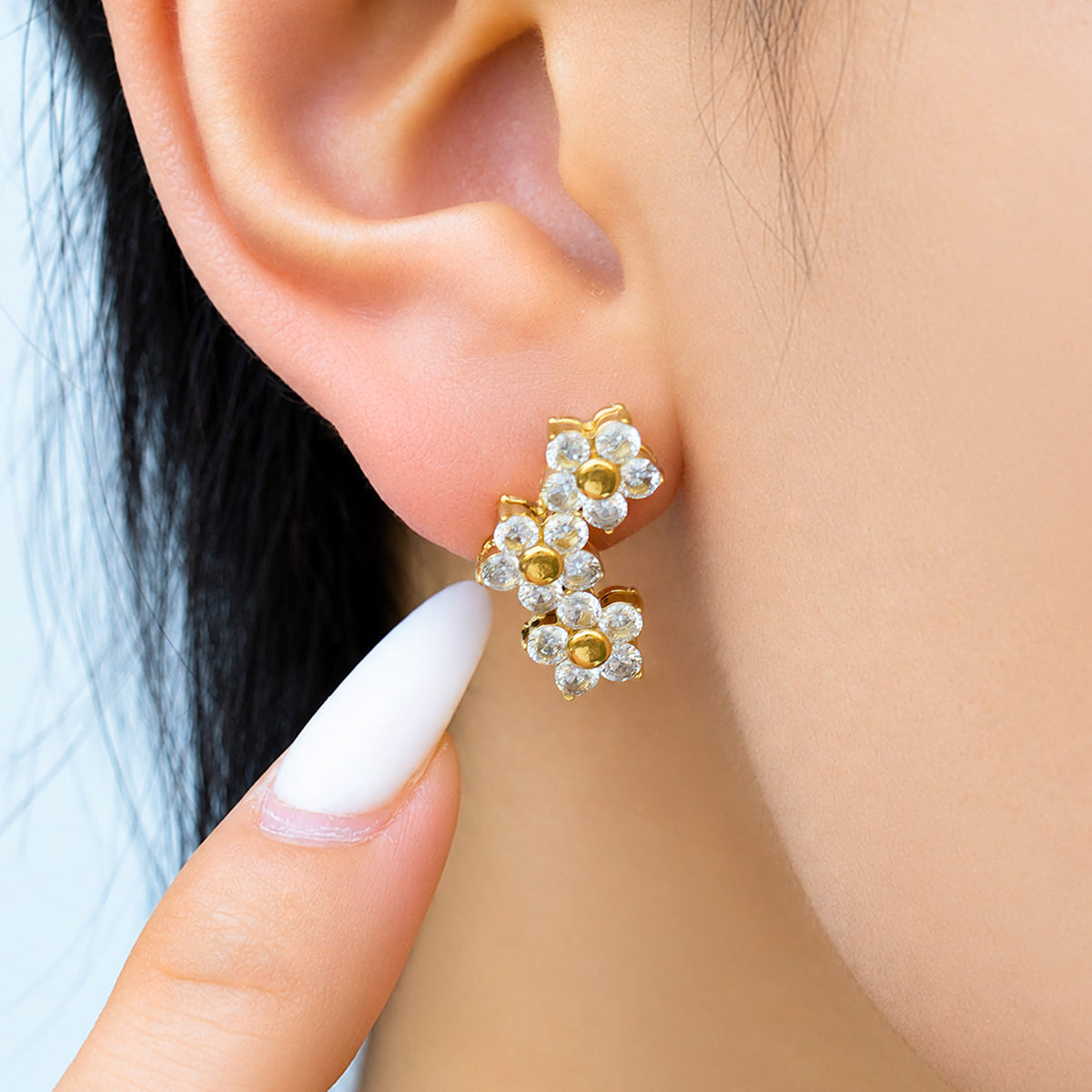 Model pointing to a gold floral drop earring featuring three connected flowers with sparkling clear stone accents
