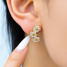 Model pointing to a gold floral drop earring featuring three connected flowers with sparkling clear stone accents