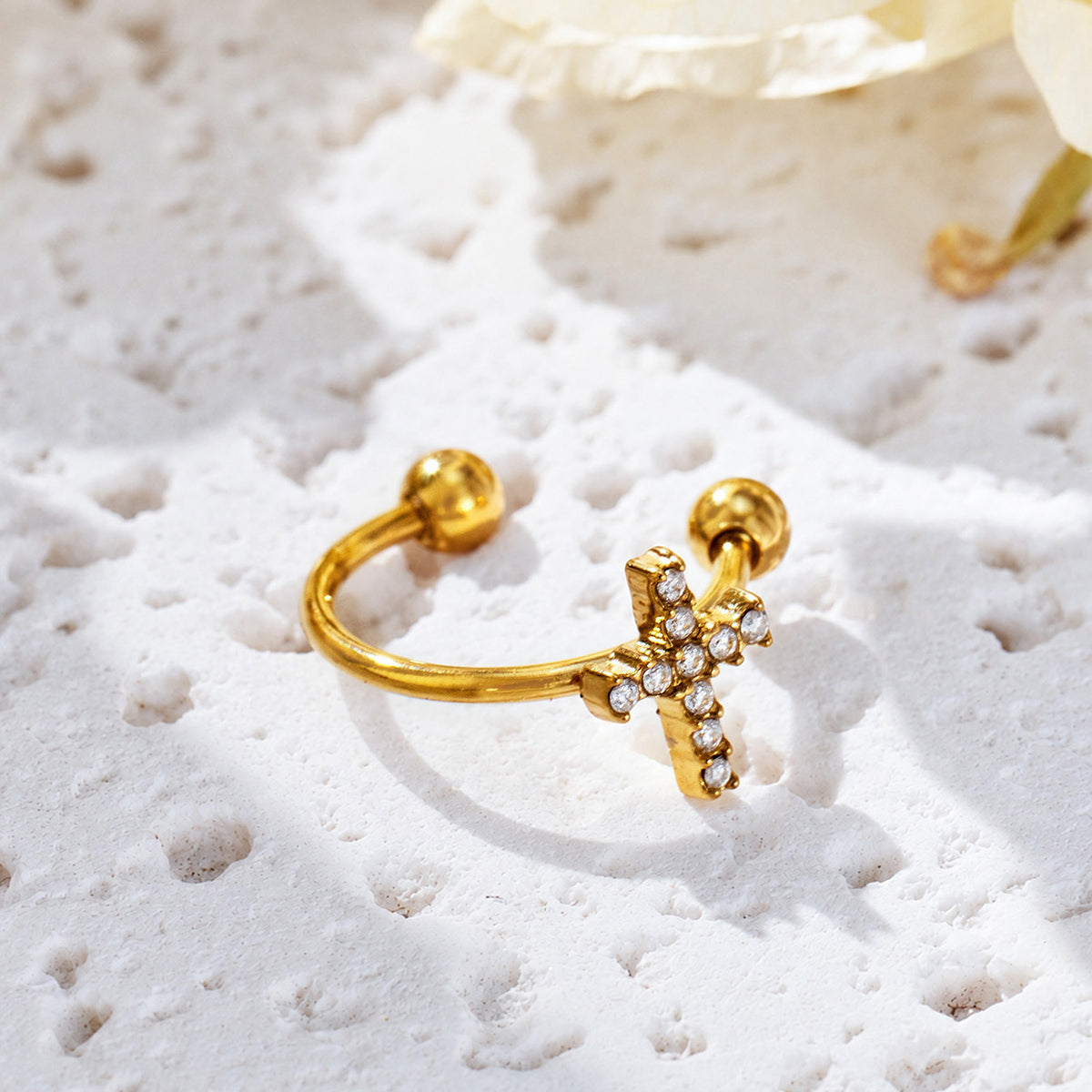 Gold-tone C-shaped ear cuff with a clear-stone cross motif resting on a textured white stone surface.