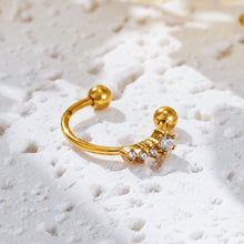 Gold-tone open ear cuff with a row of graduated clear stones along the lower curve, displayed on textured white stone.
