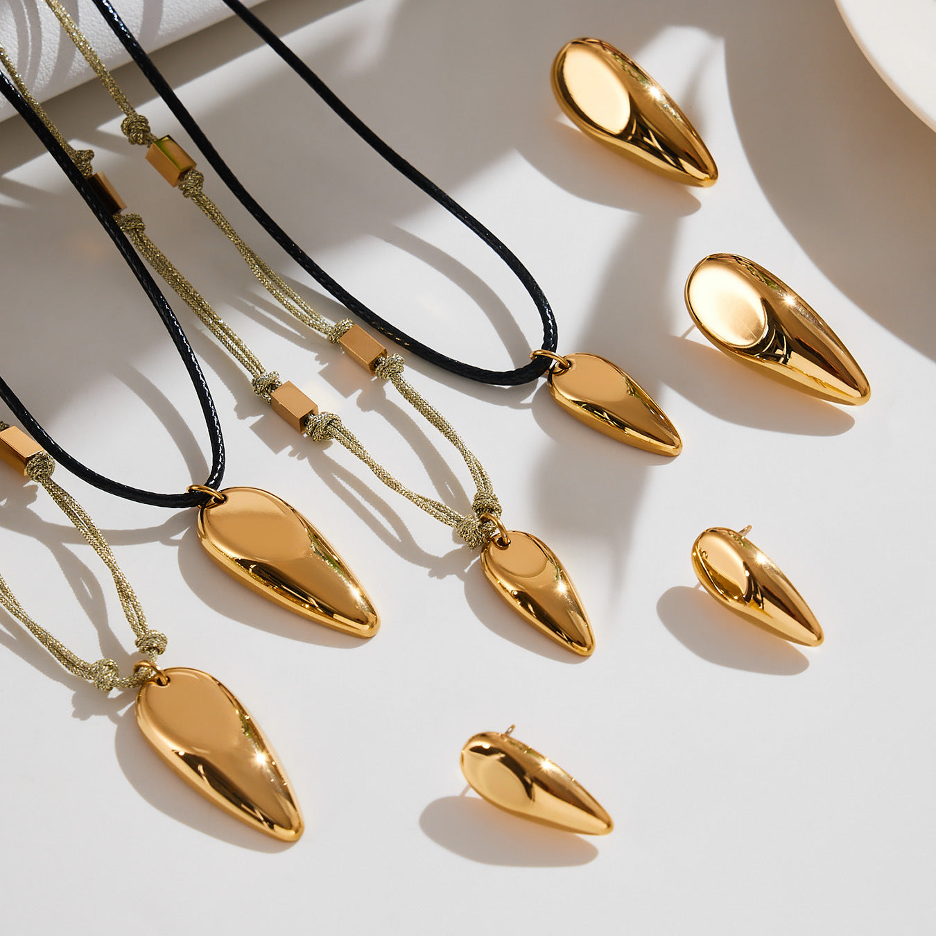 Flatlay of gold mirror-finish teardrop earrings and cord pendant necklaces made of 316L stainless steel with studio light and shadows