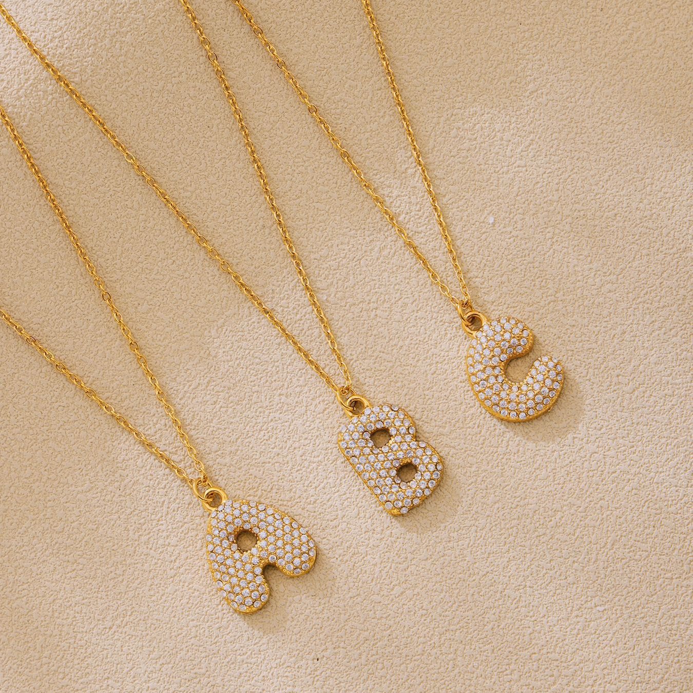 Gold A B and C iced out initial letter necklaces with zircon pave pendants on beige flatlay background