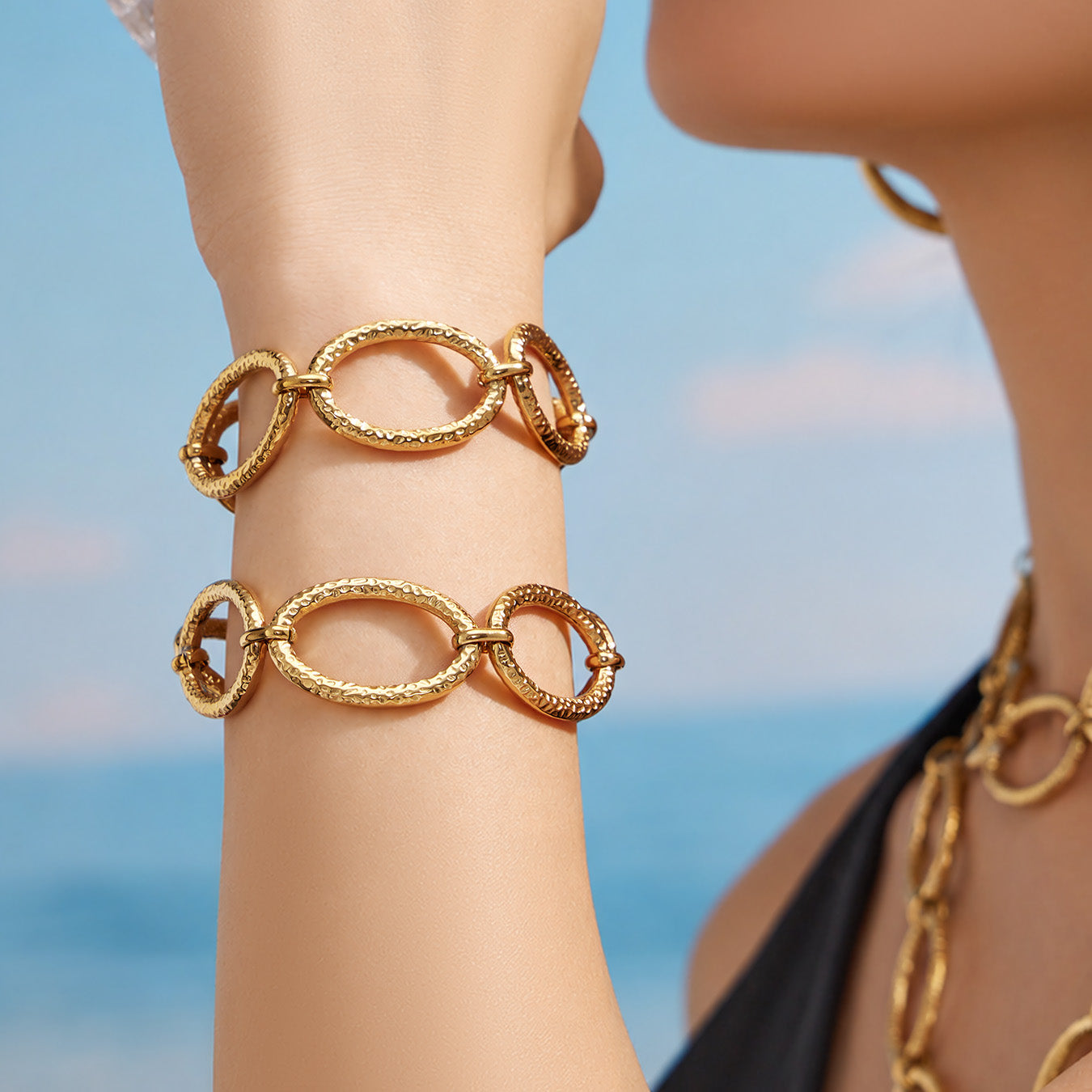 Gold chunky oval link bracelet worn on the wrist against a seaside background