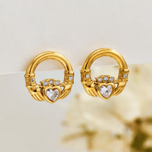 Gold Claddagh-inspired earrings displayed on a light neutral surface with clear stone hearts and a softly blurred floral background