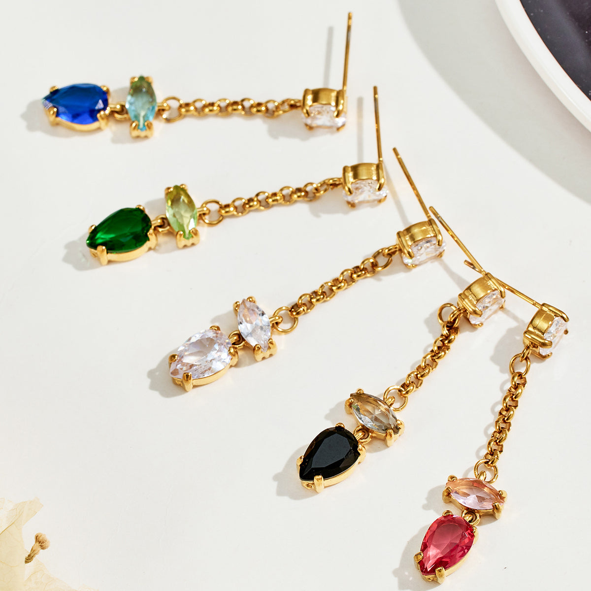 Flat lay of gold chain drop earrings featuring blue, green, white, black and red teardrop stones on a light surface