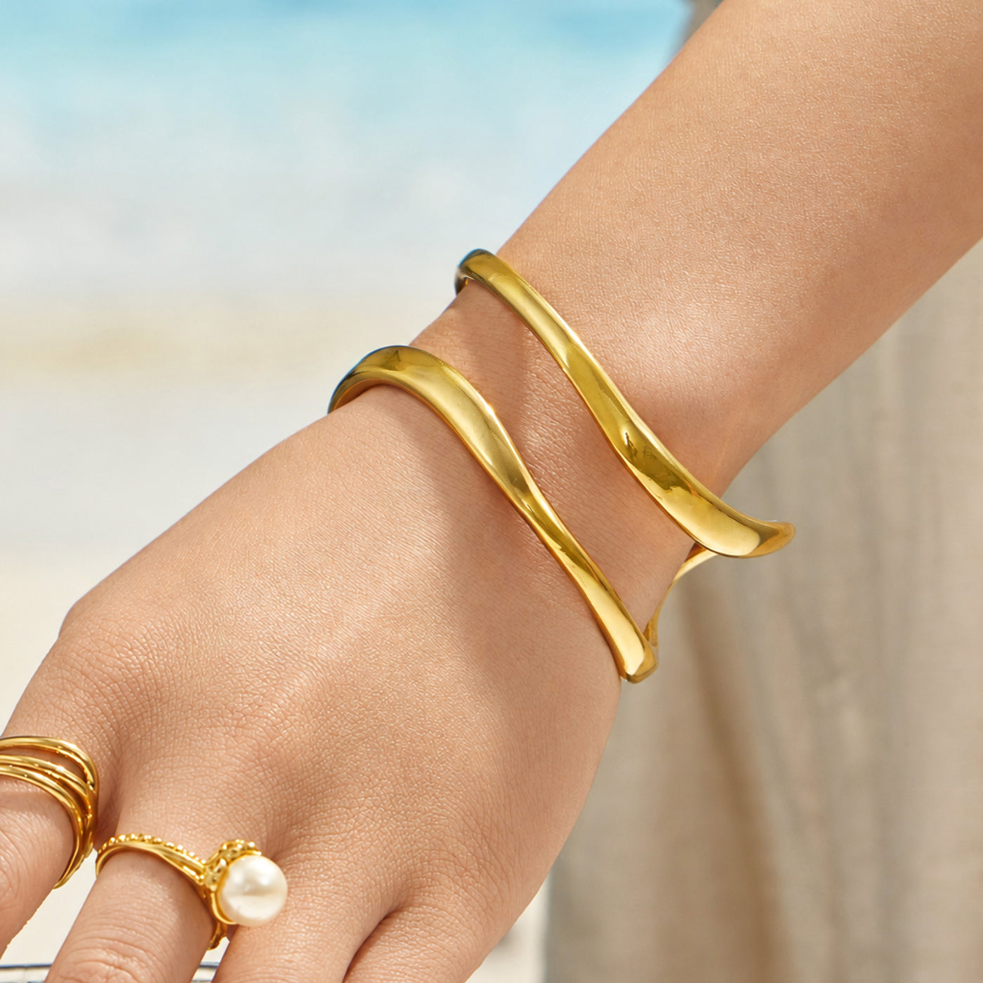 Gold double loop cuff bracelet worn on a model wrist against a blue seaside background