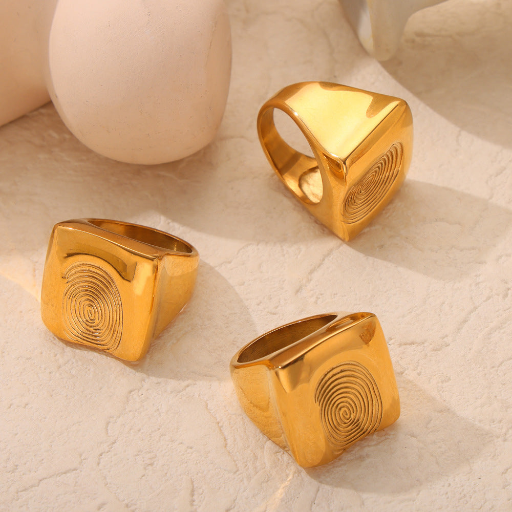 Close-up of a gold-tone fingerprint statement ring with wide square face and concentric texture