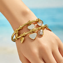 Gold heart charm bangle bracelet worn on a model wrist against a blue background