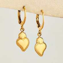 Pair of gold heart charm huggie earrings displayed on a neutral jewelry stand