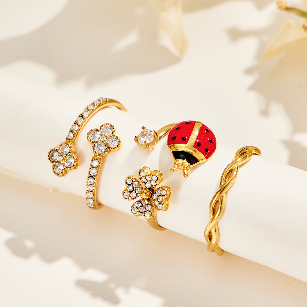 Gold-tone stainless steel ladybug, double-flower and twisted band rings arranged along a cream display ledge