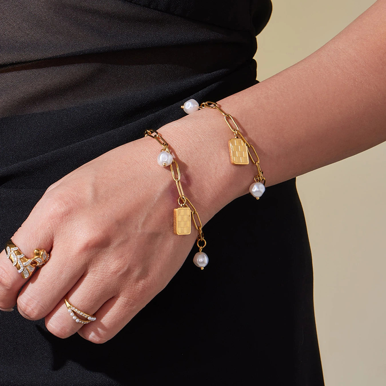 Gold paperclip charm bracelets with checkerboard and pearl-look charms worn on wrist