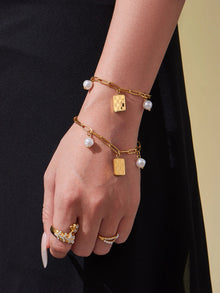 Two gold paperclip charm bracelets with pearl-look beads and checkerboard pendants on hand