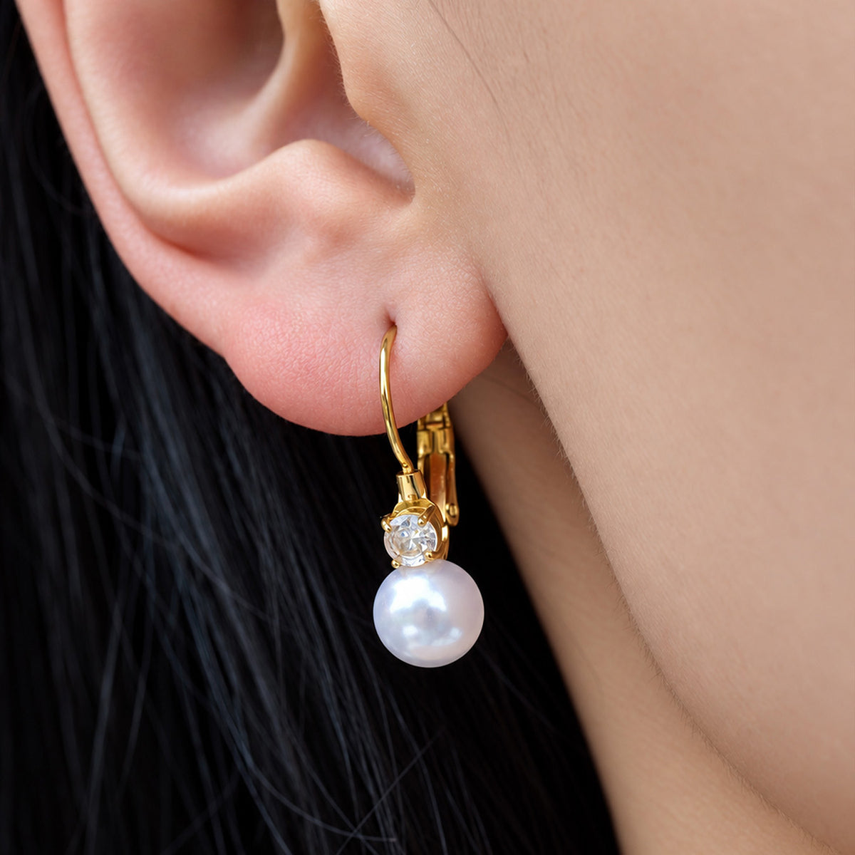 Close-up of a gold-tone pearl drop earring featuring a glossy white shell pearl suspended below a sparkling stone