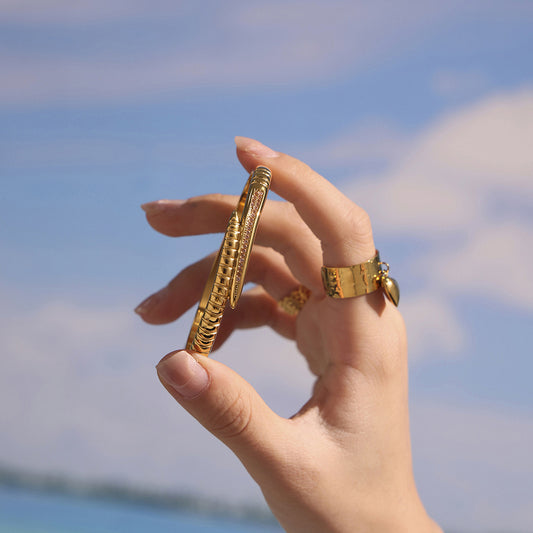 Hand holding a gold-tone nail bangle bracelet with pink zircon accents against a blue sky