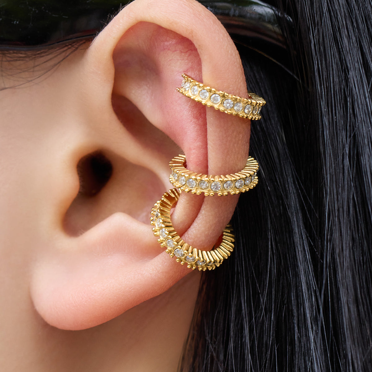 Side view of a model wearing three gold stainless steel ear cuffs with sparkling stones and ribbed details