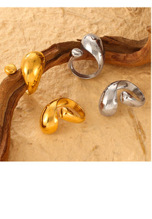 Rounded asymmetric open statement rings in gold-tone and steel-tone finishes on a textured surface