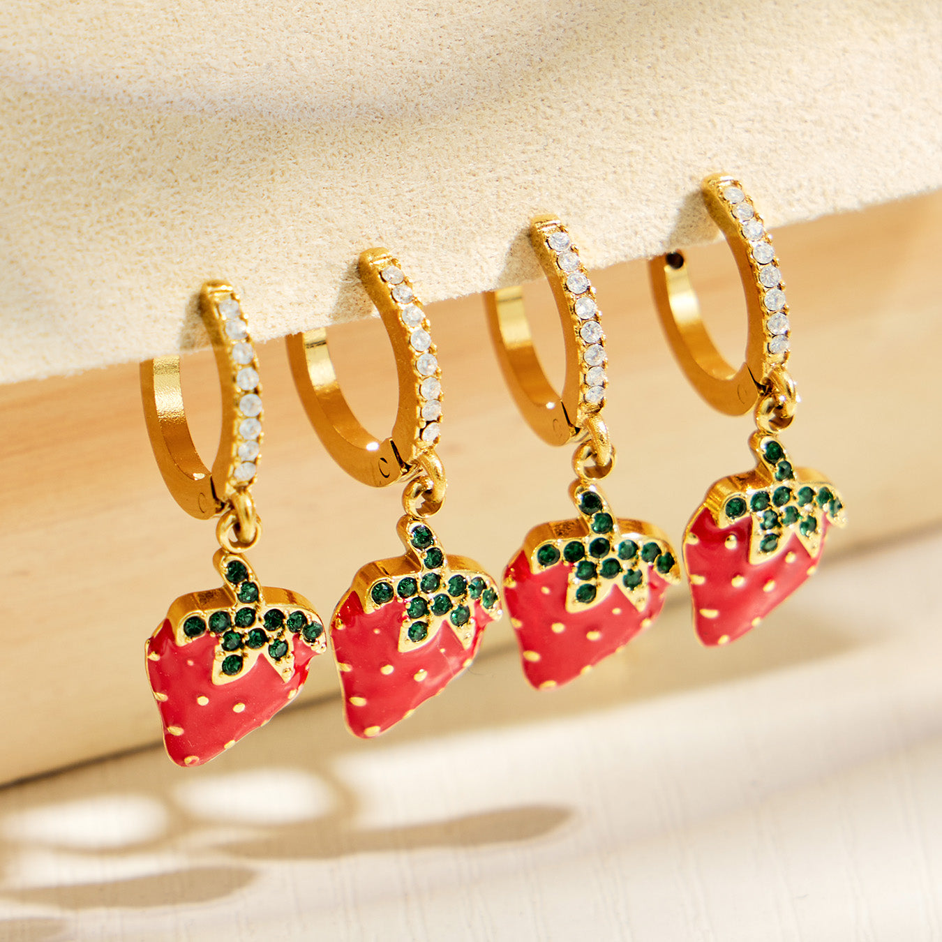 Row of gold strawberry charm huggie earrings showing the colorful fruit design