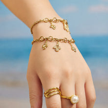 Gold teardrop charm bracelets worn together on the wrist against a beach background