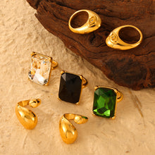 Gold-tone sculptural rings and rectangular clear, black and green stone rings beside a wooden display