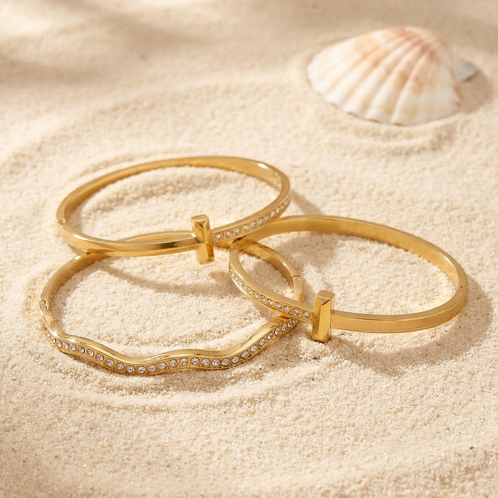 Gold zircon bangle bracelet set displayed on beach sand with shell, featuring H buckle cuff and wavy zircon bracelet designs