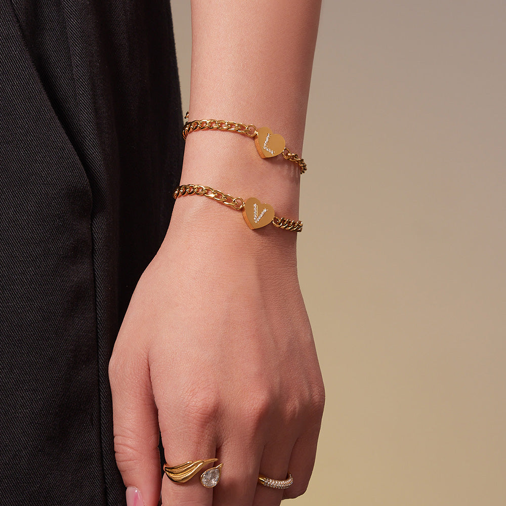 Model wearing layered gold-tone initial heart bracelets with a black outfit